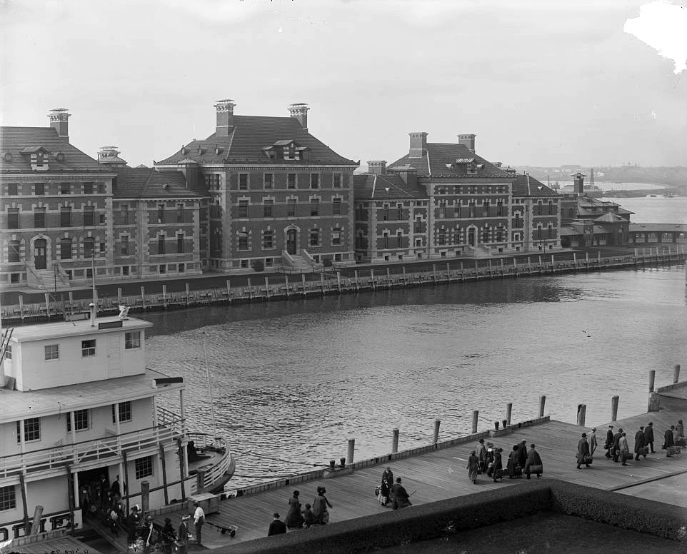 Black and white photograph of immigrants walking across a boardwalk at Ellis Island