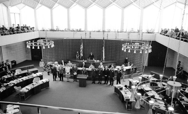 Senate floor. Members of the congress stand around desks in the front and sides of the room. Two chandeliers hang over an open forum in the middle.