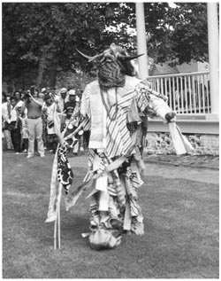 A performer is dressed in several types of cloths covering their entire body and a horned hat or helmet on their head. There is a crowd of people standing behind watching and man with a large camera. 