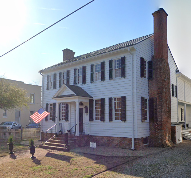 A white, Federal-style house. It is a clear day. A brick chimney, a waving American flag, and some nice landscaping mark the property. 