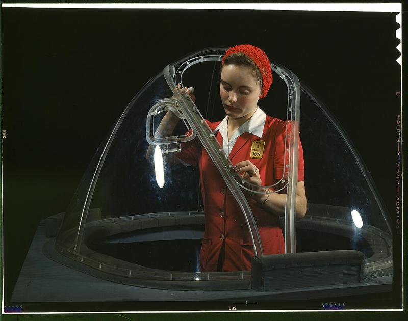 Woman working on a bomber cockpit. She is wearing red and has a red headwrap. 