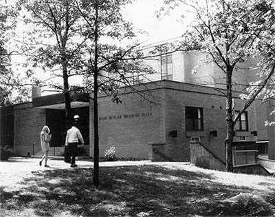 A brick building with a few nearby trees. Some students are walking towards the building.